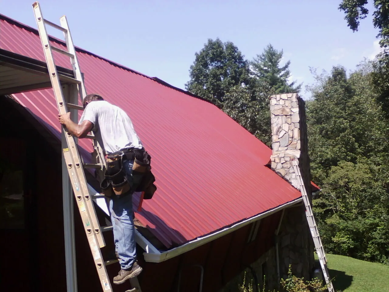 Roofer on a ladder beside a red metal roof and stone chimney — Soffit & Fascia Repair by Roofing Brothers in Aledo